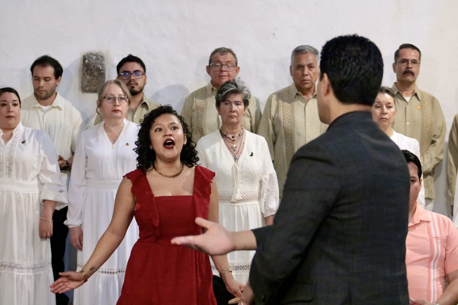 Natali Juárez en vestido rojo cantando con coro detrás y Omar Nieto dirigiendo
