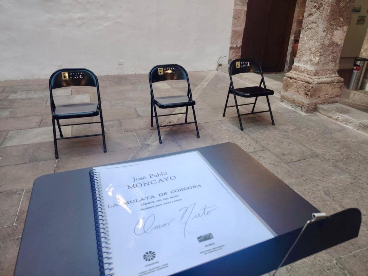 Partitura de La Mulata de Córdoba firmada por Omar Nieto, sobre atril con sillas de Ópera de Morelia en el patio del Museo del Estado